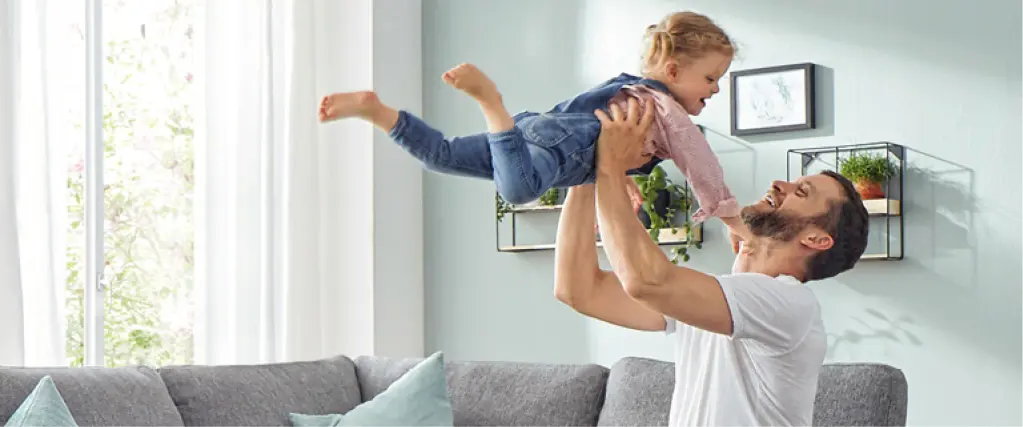 Vater hebt lachendes Kind in die Luft vor grauem Sofa im hellen Wohnzimmer. Familienmoment mit moderner Wohnzimmereinrichtung und gemütlicher Wohnlandschaft